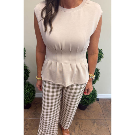 A woman modeling a sleeveless beige top with a structured corset-style waist and pleated peplum hem, paired with brown and beige gingham wide-leg pants, standing indoors.