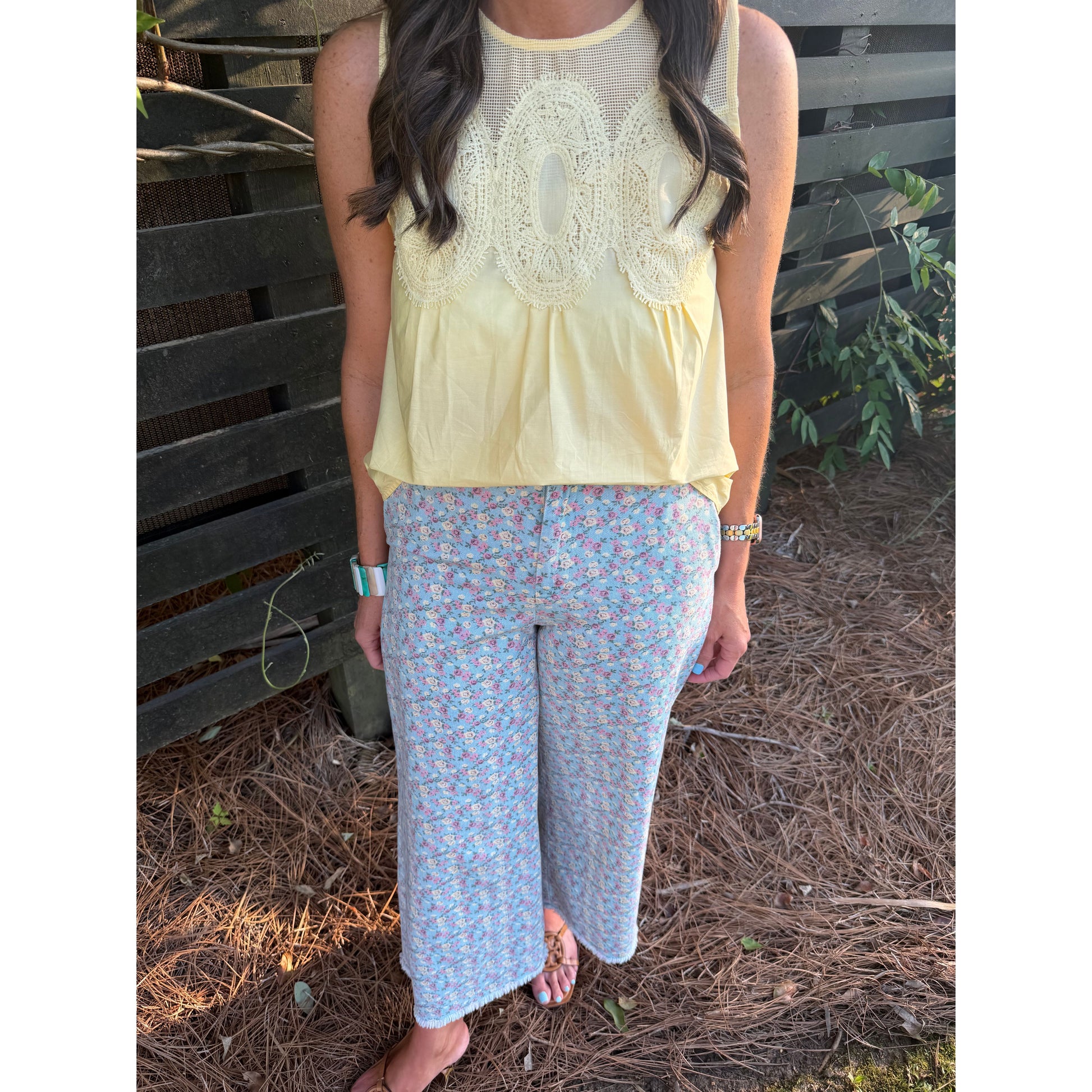 Woman wearing a light yellow top and floral pants standing against a wooden fence.