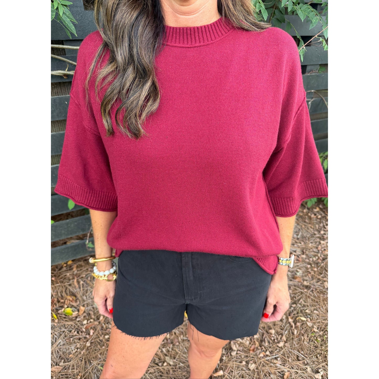 A woman wearing a ribbed crewneck knit sweater in deep burgundy with elbow-length sleeves, paired with black shorts, standing outdoors in front of a dark wooden fence.