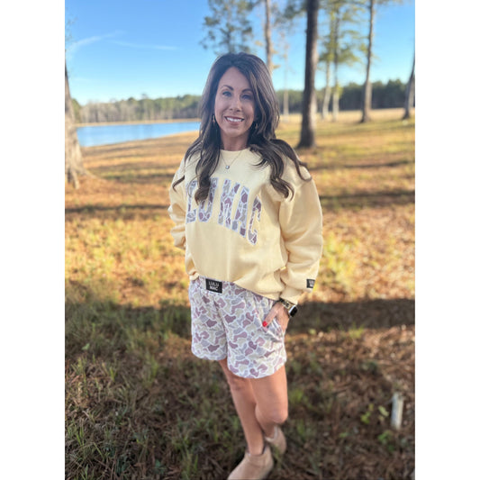 Woman wearing a light-colored sweatshirt and patterned shorts standing in a grassy field with trees and a lake in the background.