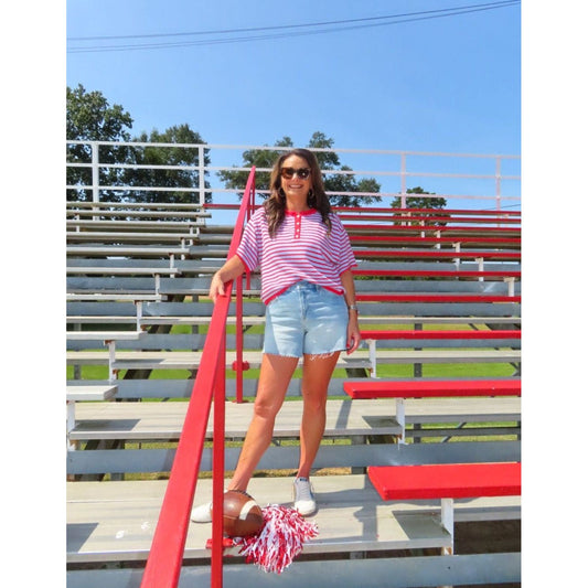 A woman wearing a pink and white striped polo shirt, denim shorts, and white sneakers stands on red and gray metal bleachers outdoors under a blue sky, holding a pink and white pom-pom.