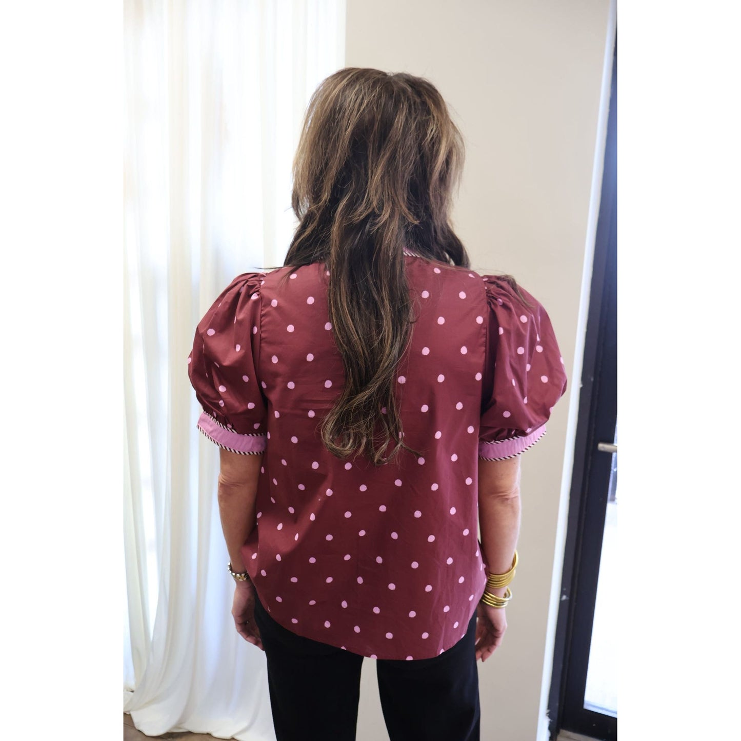 A woman viewed from behind wearing a burgundy short-sleeve blouse with pink polka dots and puff sleeves, paired with black pants.