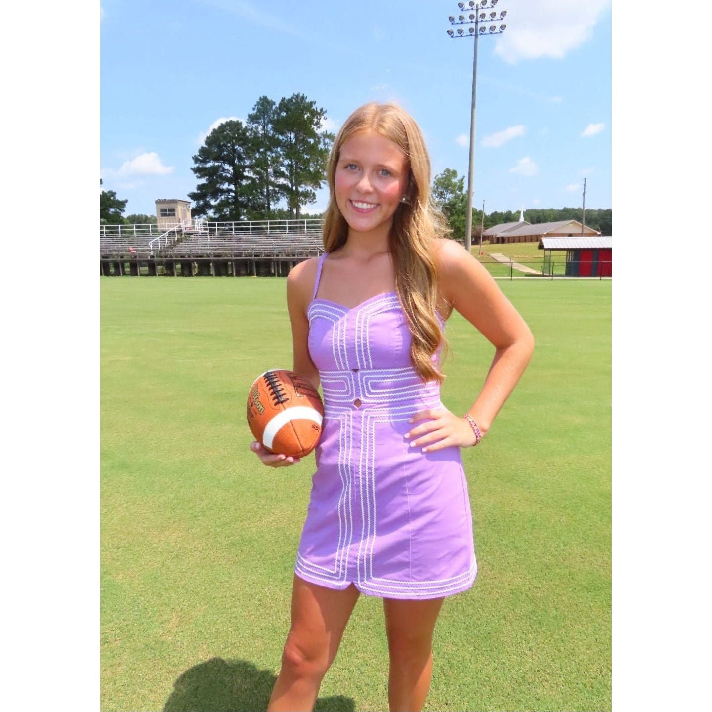 A woman stands on a grassy sports field holding a football, wearing a lavender sleeveless mini dress with white geometric stripe patterns. The background shows bleachers, trees, and a blue sky.