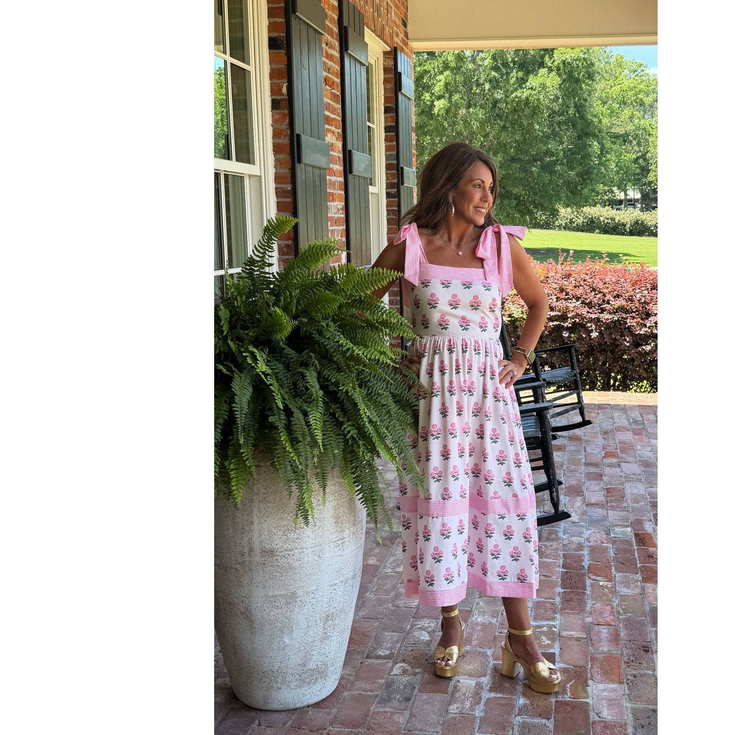 Woman in a pink floral dress standing on a patio with greenery and a house in the background