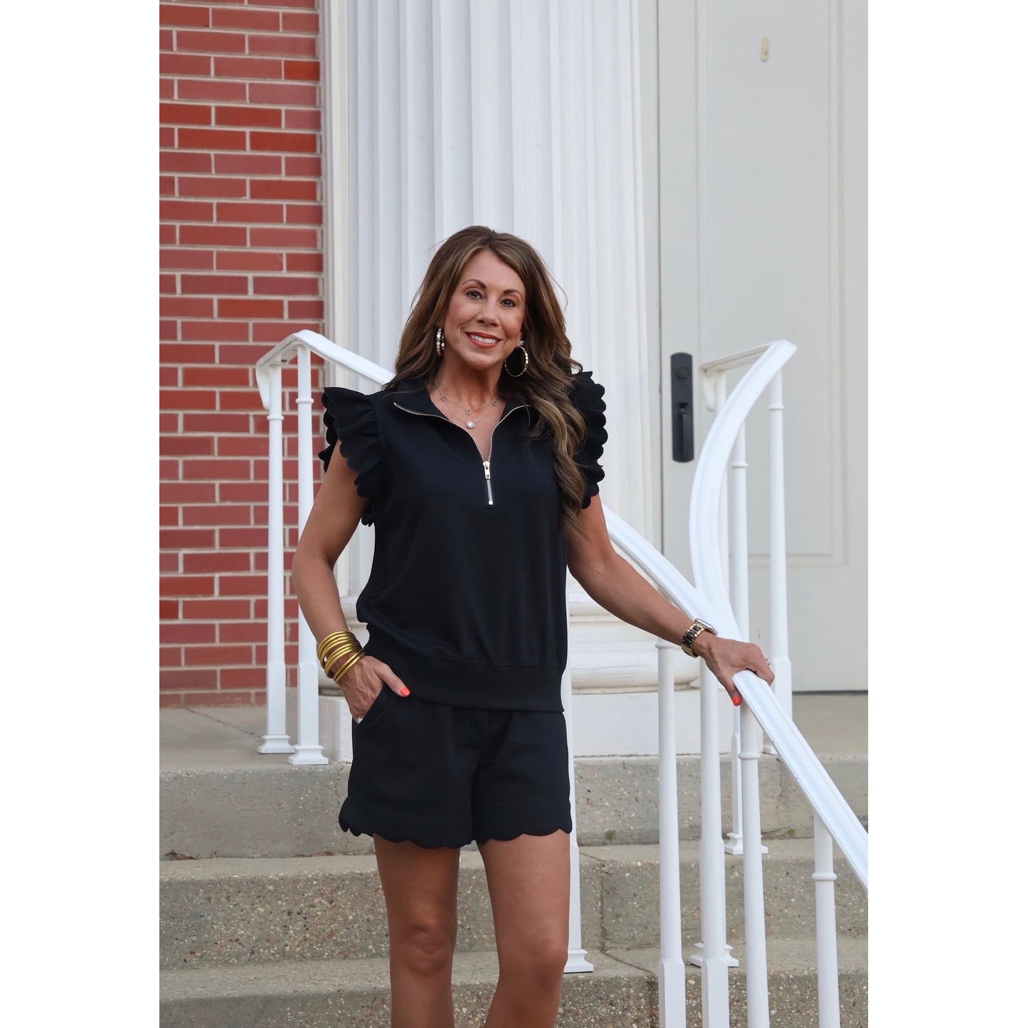 Woman in black outfit standing on steps with a white railing and brick wall background