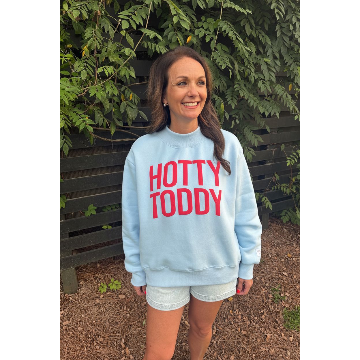 A woman wearing a light blue crewneck sweatshirt with 'HOTTY TODDY' printed in red across the chest, standing outdoors in front of a wooden fence and green foliage.
