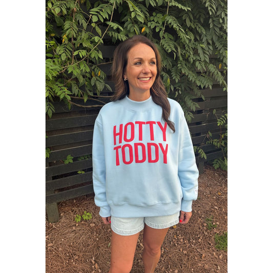 A woman wearing a light blue crewneck sweatshirt with 'HOTTY TODDY' printed in red across the chest, standing outdoors in front of a wooden fence and green foliage.