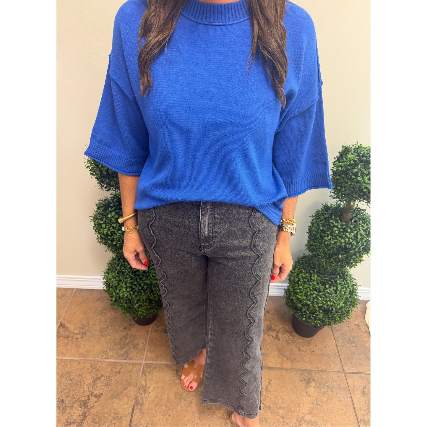 A woman wearing a vibrant royal blue knit sweater with elbow-length sleeves and dark gray scalloped wide-leg jeans, standing in front of a beige wall with decorative green plants.