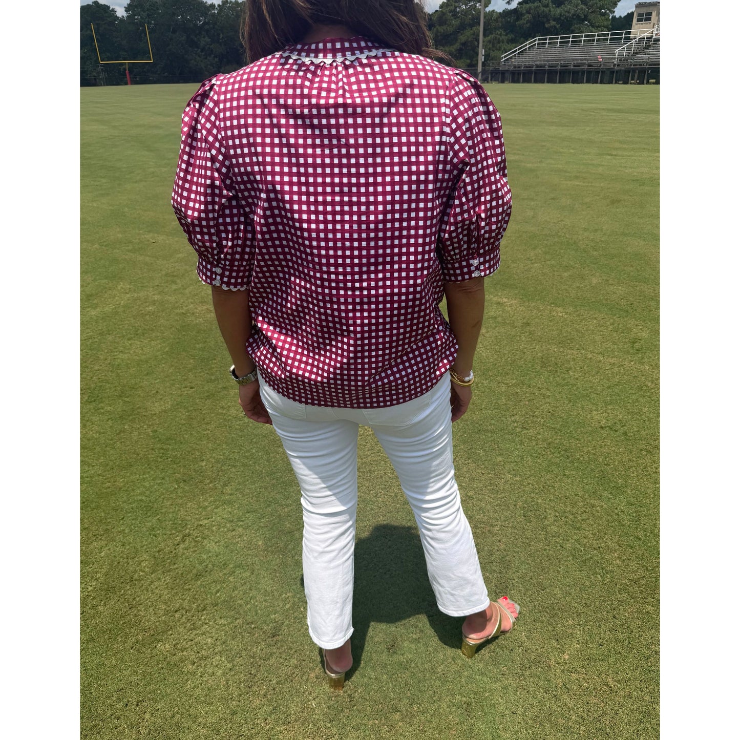 A person standing on a green grass field wearing a burgundy and white gingham short-sleeve button-up shirt, white pants, and brown sandals.