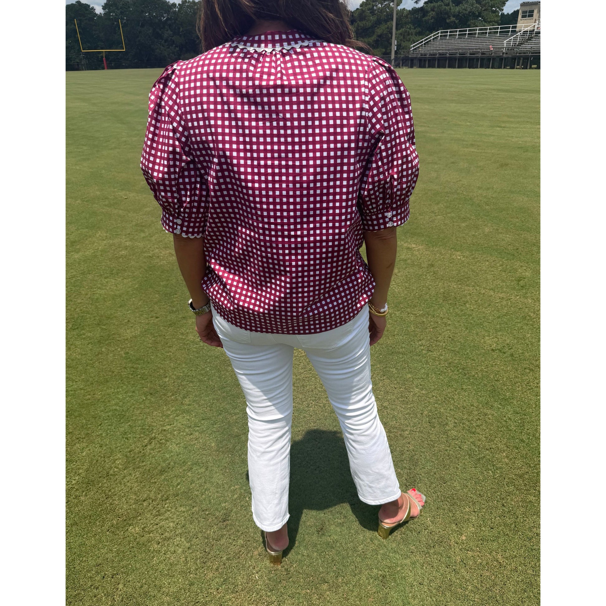 A person standing on a green grass field wearing a burgundy and white gingham short-sleeve button-up shirt, white pants, and brown sandals.