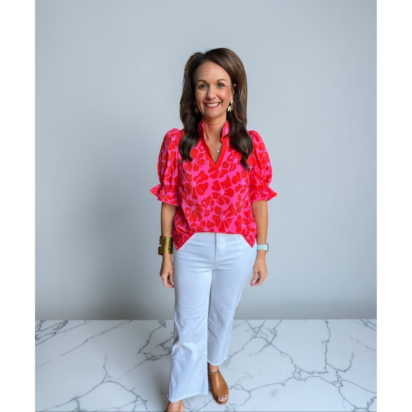 Woman wearing a bright pink blouse with a pattern and white pants on a gray background