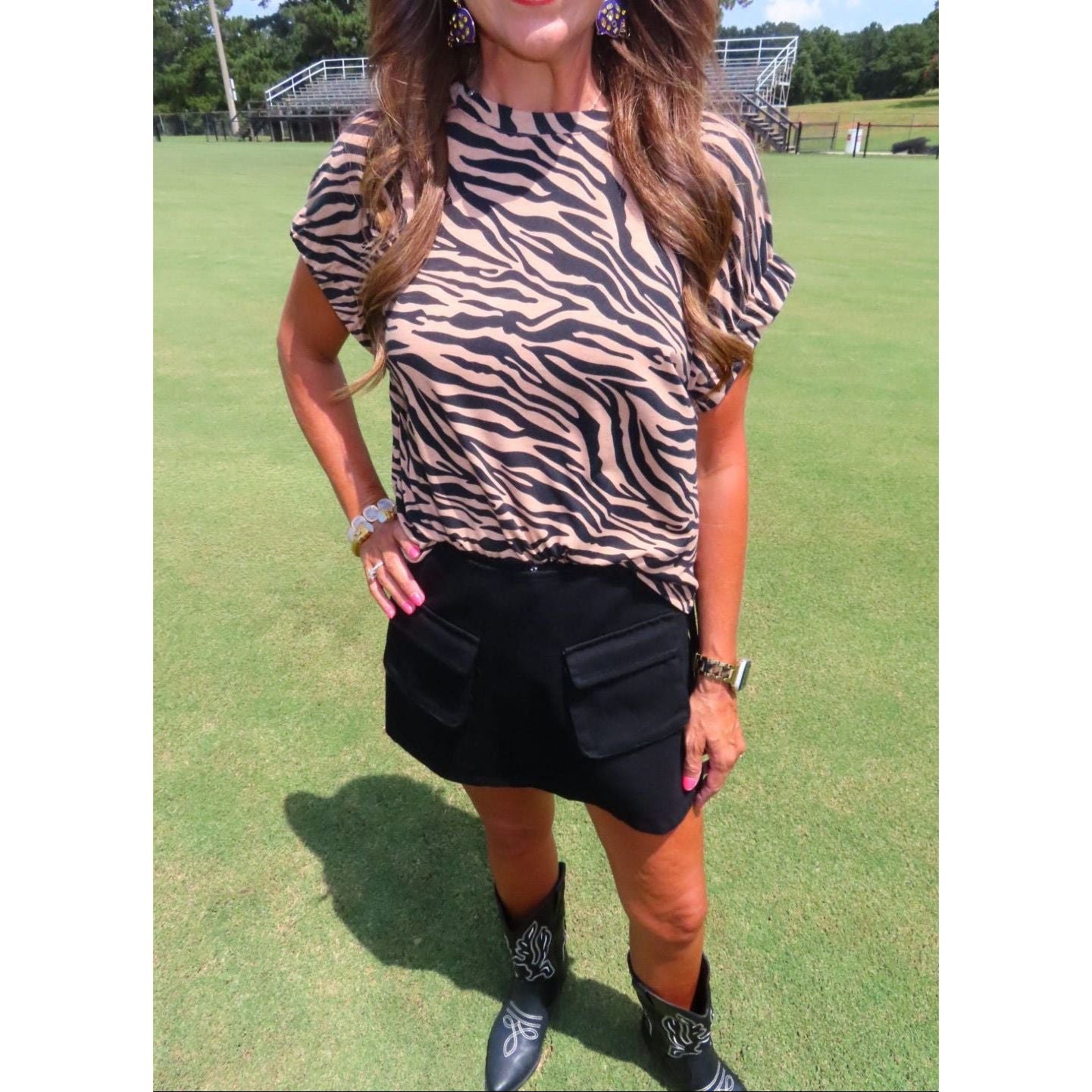 A woman wearing a beige and black zebra-print short-sleeve top and a black mini skirt, standing on a green grass field.