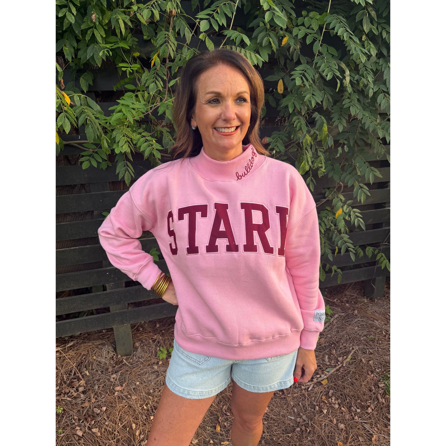 A woman wearing a pink sweatshirt with 'START' printed in maroon letters on the front, paired with light blue denim shorts, standing outdoors in front of a green leafy fence.