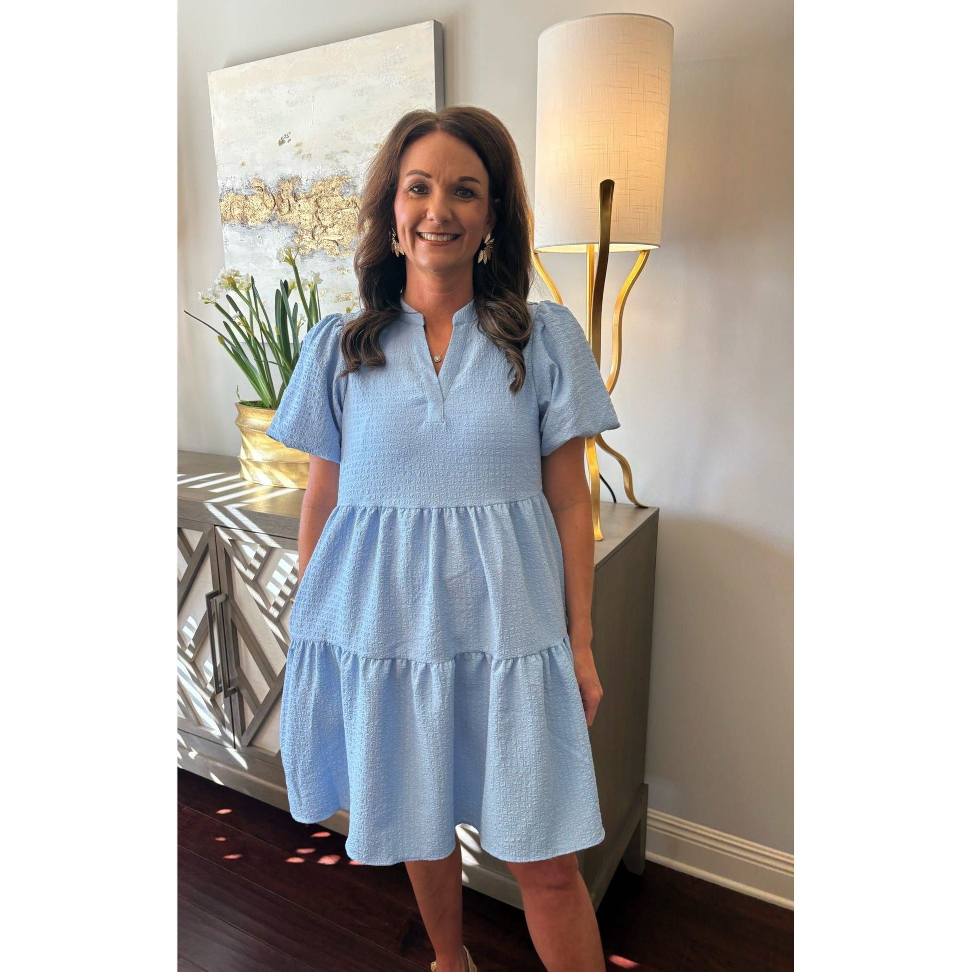 Woman in a light blue dress standing in a room with lamps and decorative items.