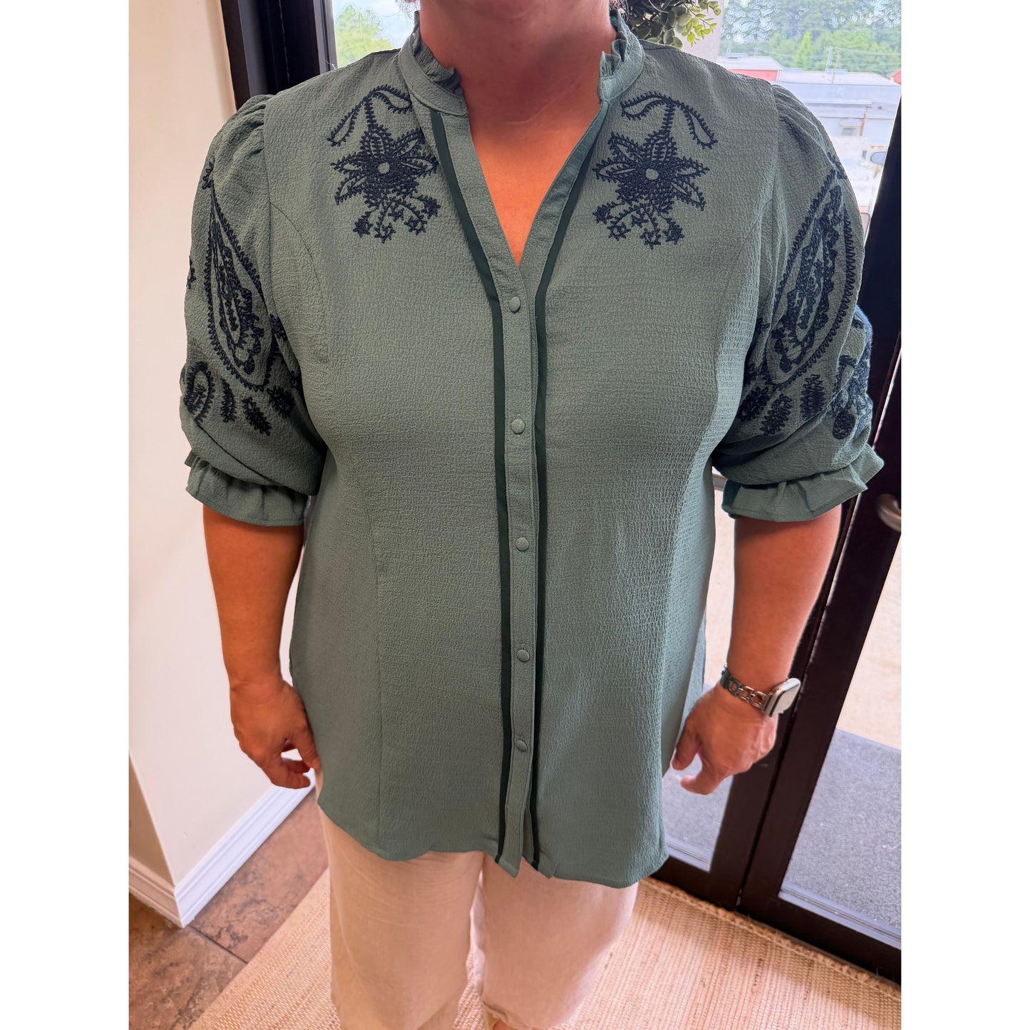 A woman wearing a sage green button-up blouse with black embroidered floral and paisley details on the shoulders and sleeves, paired with light beige pants.