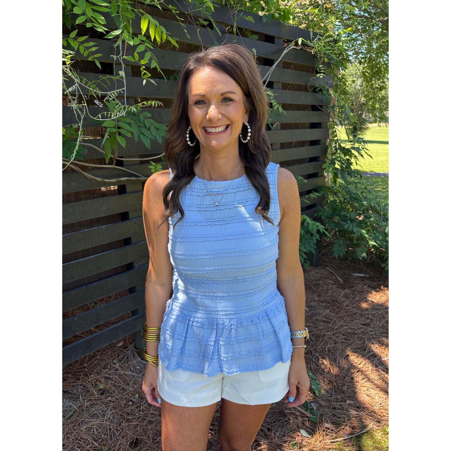 Woman in a blue top and white shorts standing outdoors with greenery around.