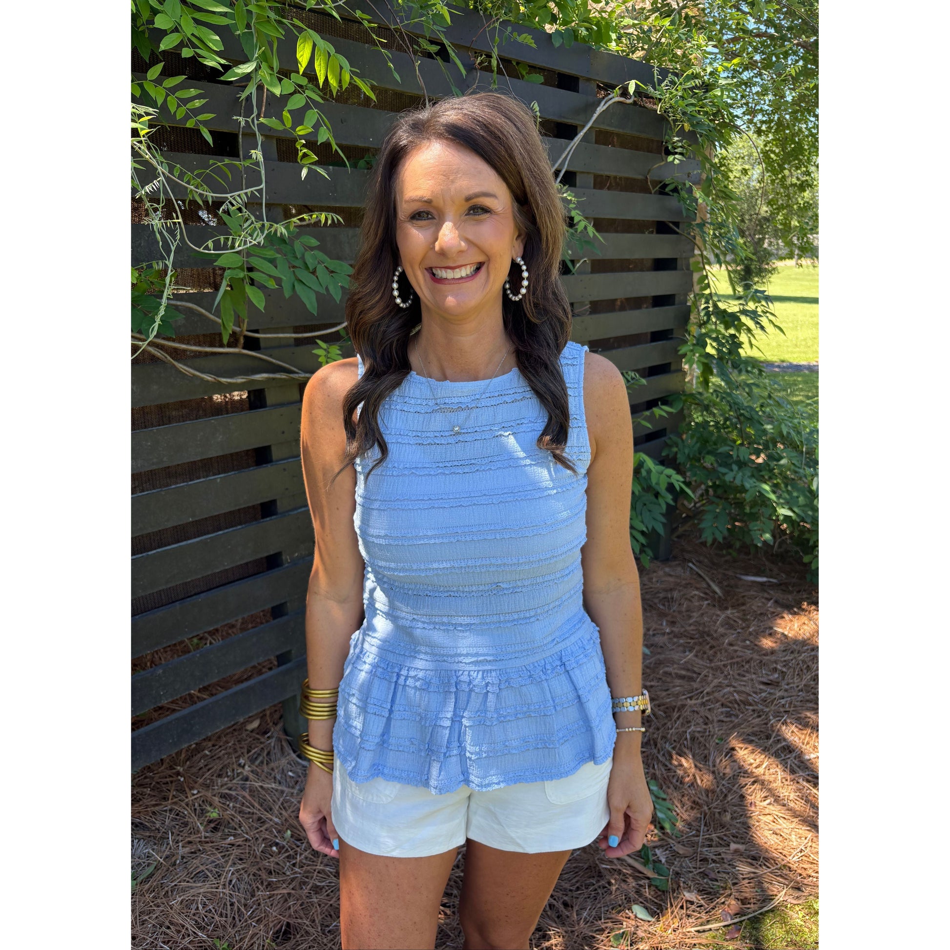 Woman in a blue top and white shorts standing outdoors with greenery around.