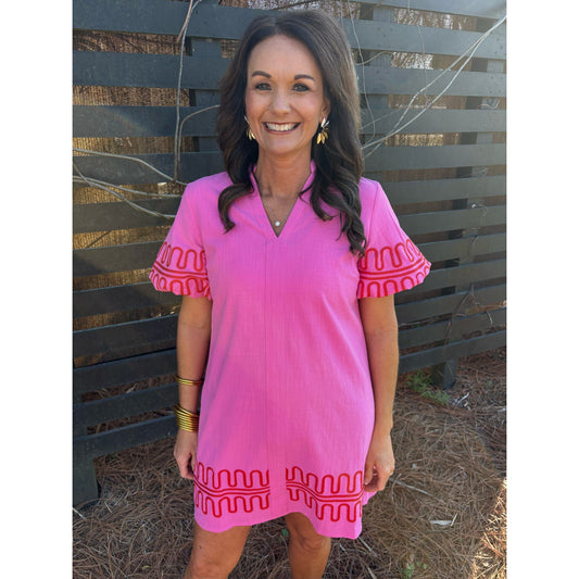 Woman wearing a pink dress with red embroidery standing in front of a wooden fence.