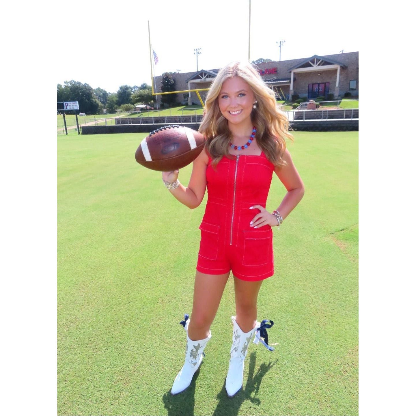 A woman stands on a grassy field holding a football, wearing a bright red sleeveless zip-front romper with matching shorts, white cowboy boots, and a blue beaded necklace.