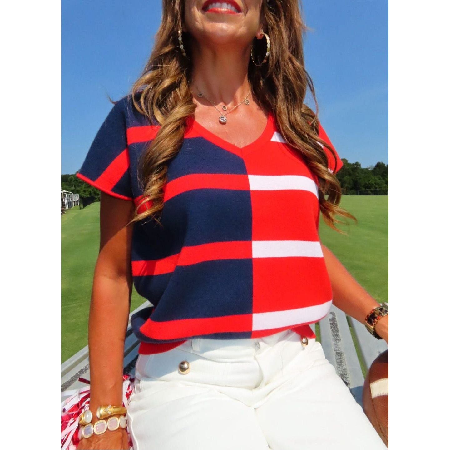 A woman wearing a red, navy, and white striped V-neck short-sleeve top and white pants, posing outdoors on a sunny day with a green golf course and blue sky in the background.