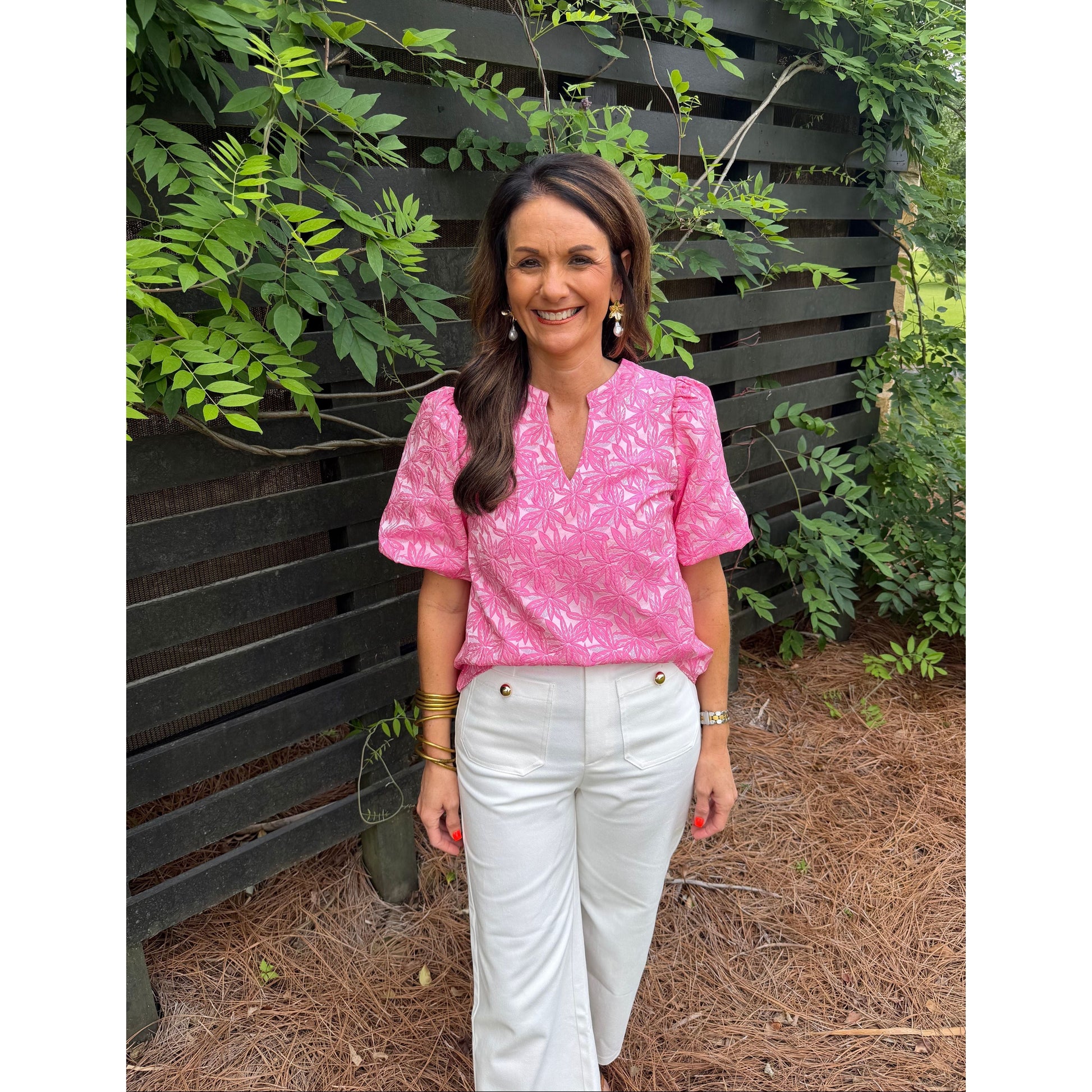 Woman in a pink blouse and white pants sitting on a black metal bench with greenery around.