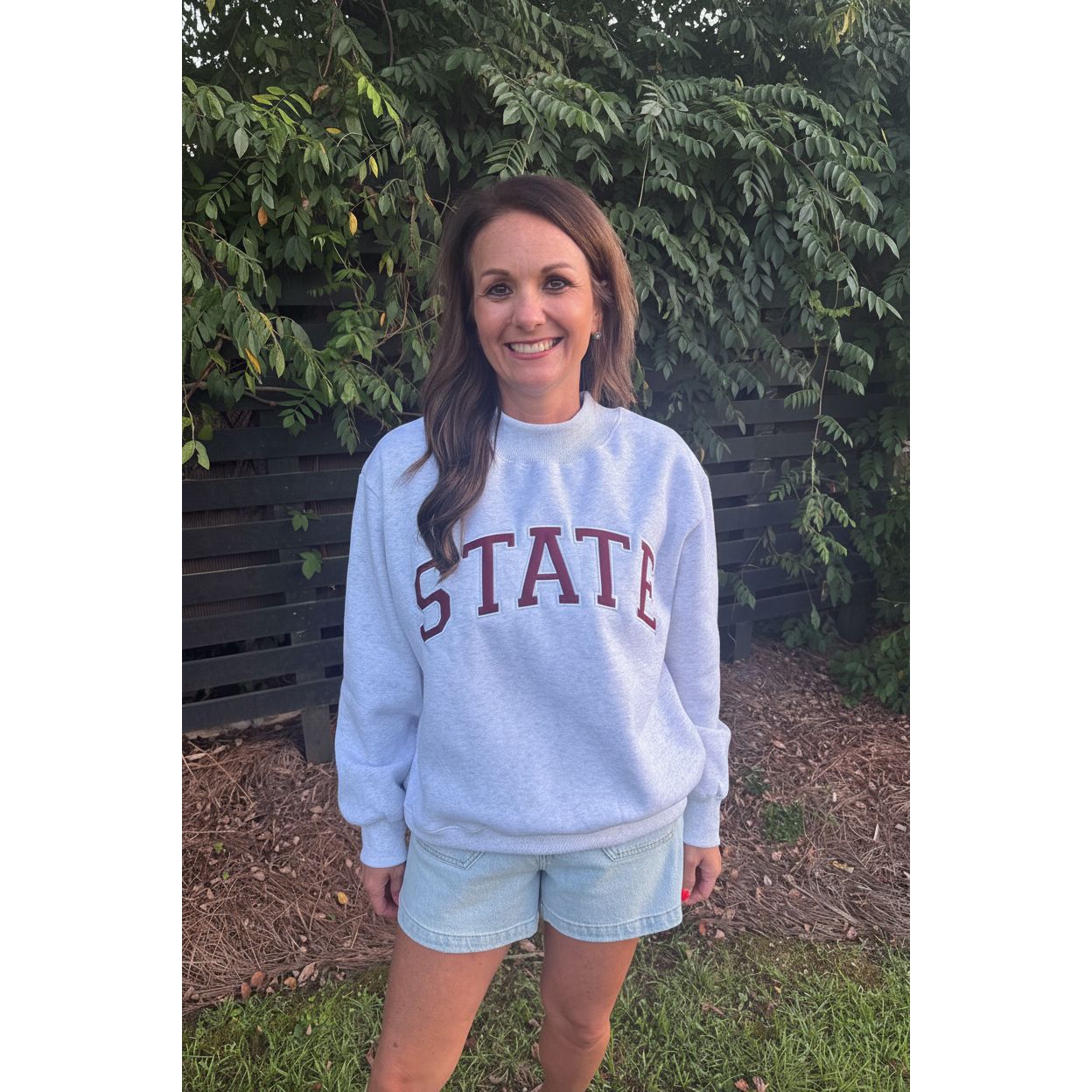 A woman wearing a light heather gray sweatshirt with 'STATE' printed in dark red across the chest, paired with light denim shorts, standing outdoors on a bench with greenery and mulch in the background.
