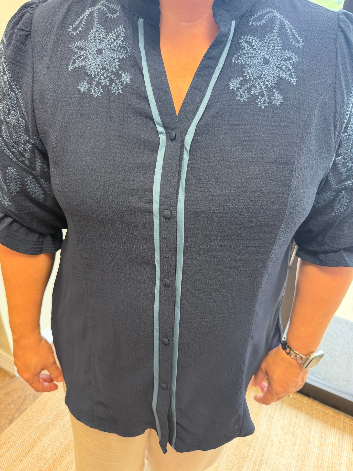A woman wearing a dark charcoal gray button-up blouse with light blue-gray embroidered floral details on the shoulders and down the front placket, paired with beige pants.