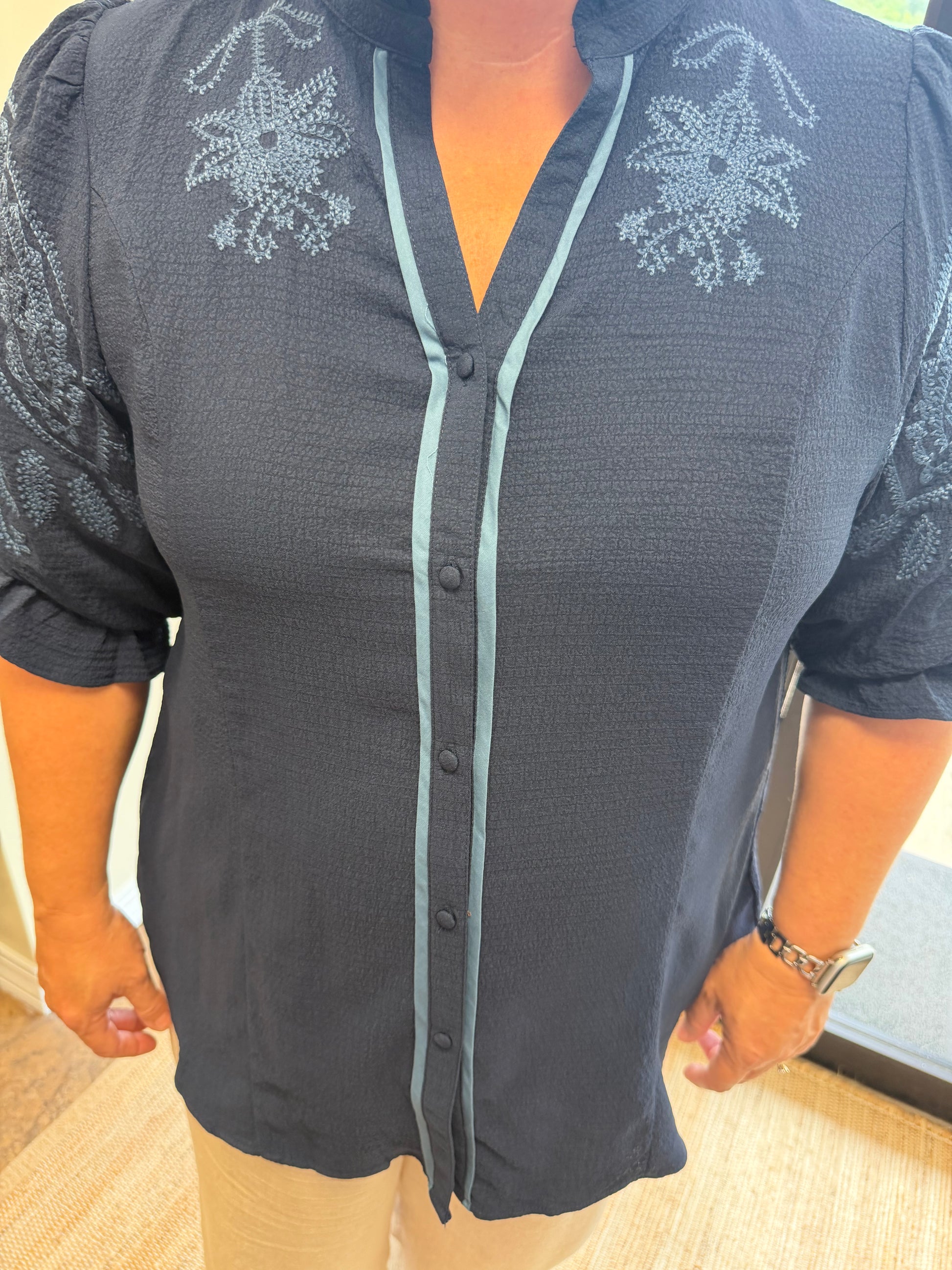 A woman wearing a dark charcoal gray button-up blouse with light blue-gray embroidered floral details on the shoulders and down the front placket, paired with beige pants.