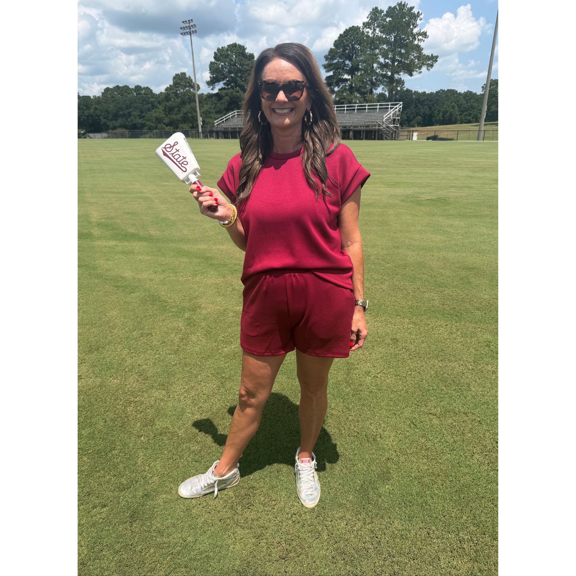 A woman wearing a matching burgundy short-sleeve top and shorts set stands on a green grass field, holding a white bottle with a red and white label.
