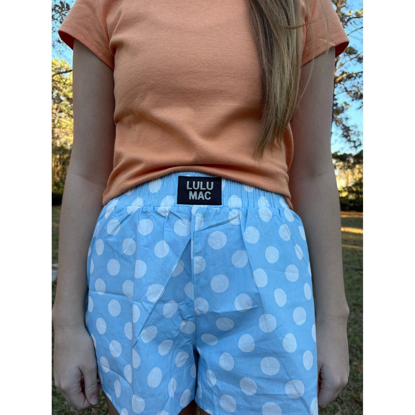 Person wearing a peach shirt and blue polka dot shorts with Lulu Mac label outdoors.