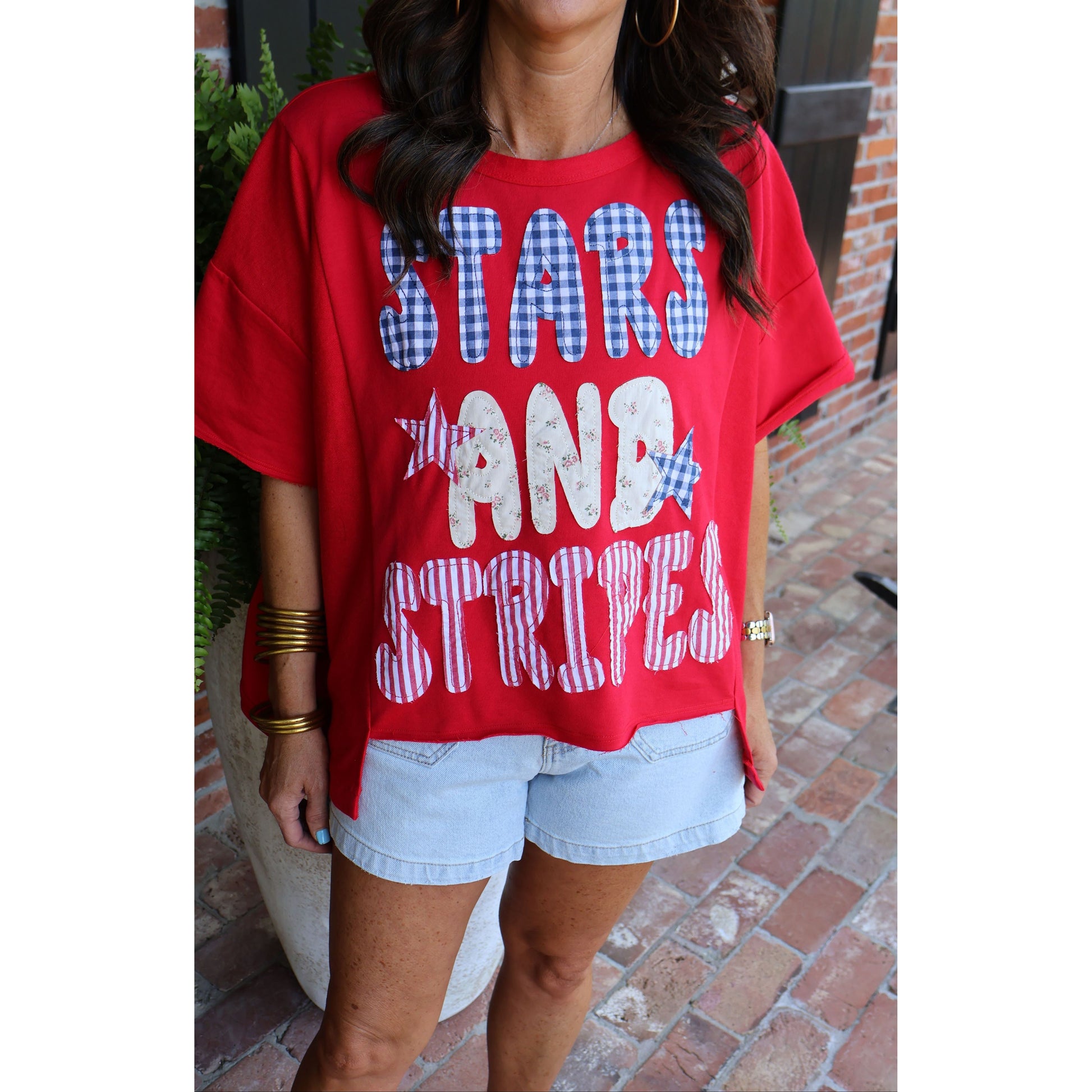 Person wearing a red t-shirt with 'STARS AND STRIPES' text, standing on a brick pavement.