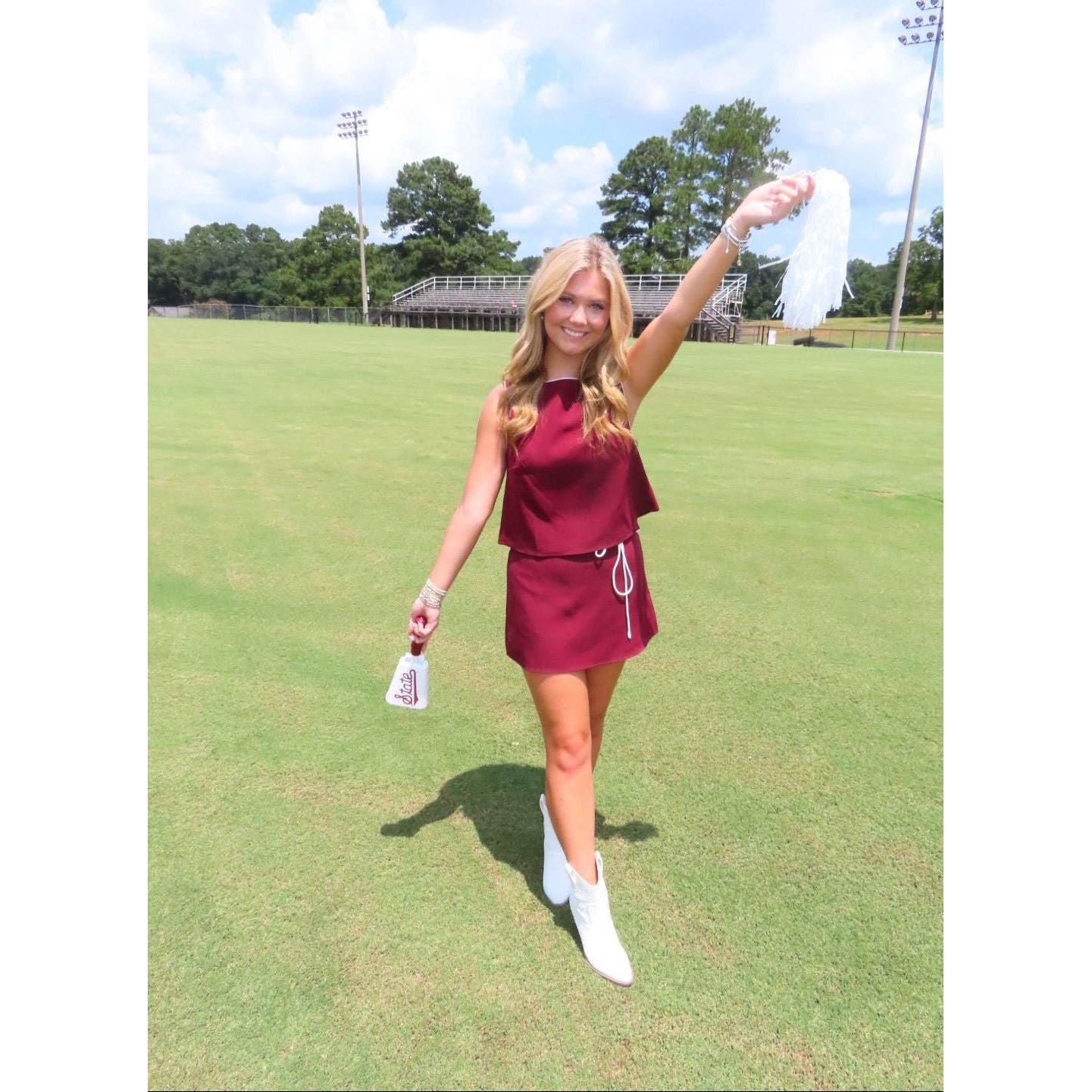 A woman stands on a grassy field under a cloudy sky, wearing a sleeveless burgundy mini dress with a white drawstring and white ankle boots, holding a white towel in one hand and a small bag in the other.