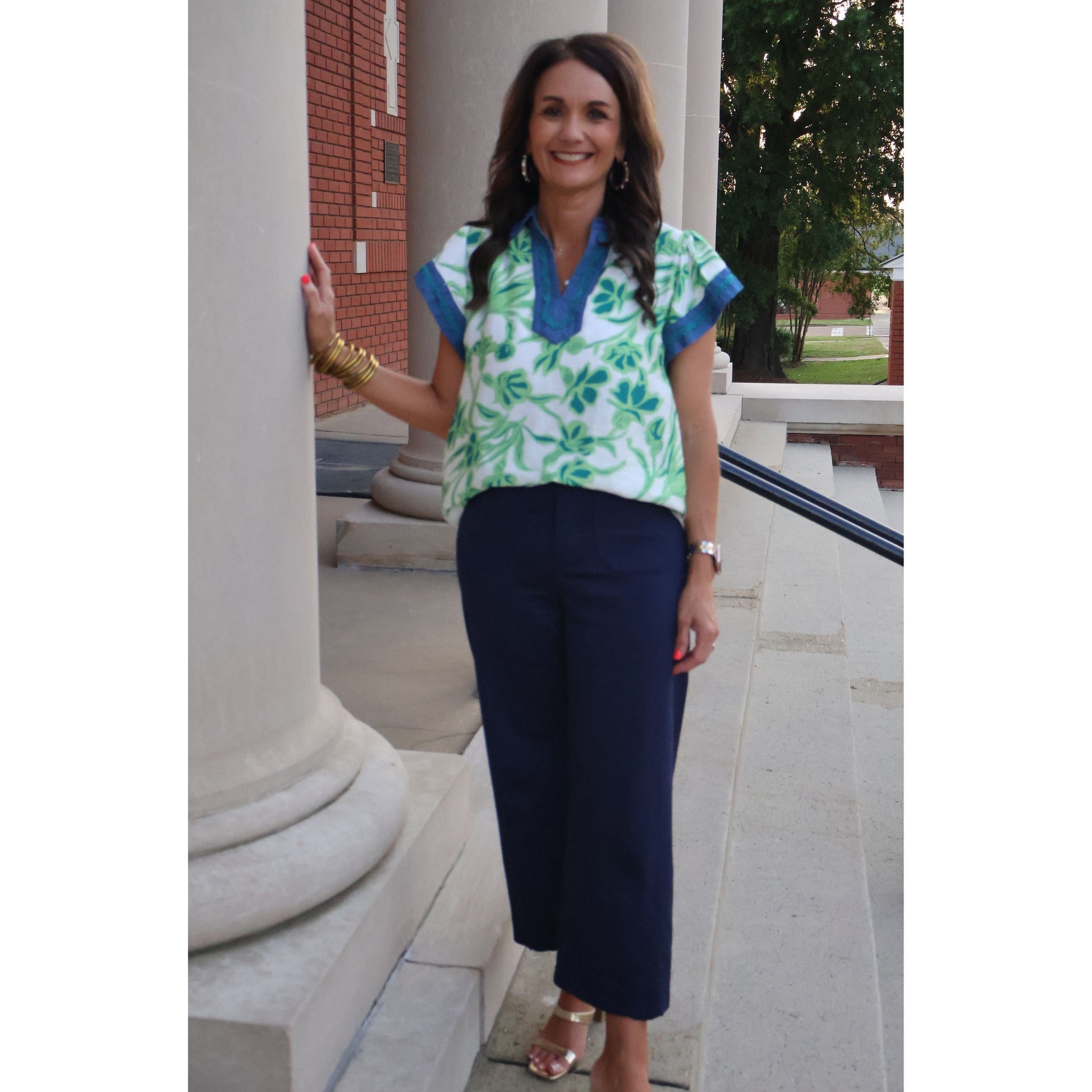 Woman in a floral blouse and navy pants standing outdoors near columns.