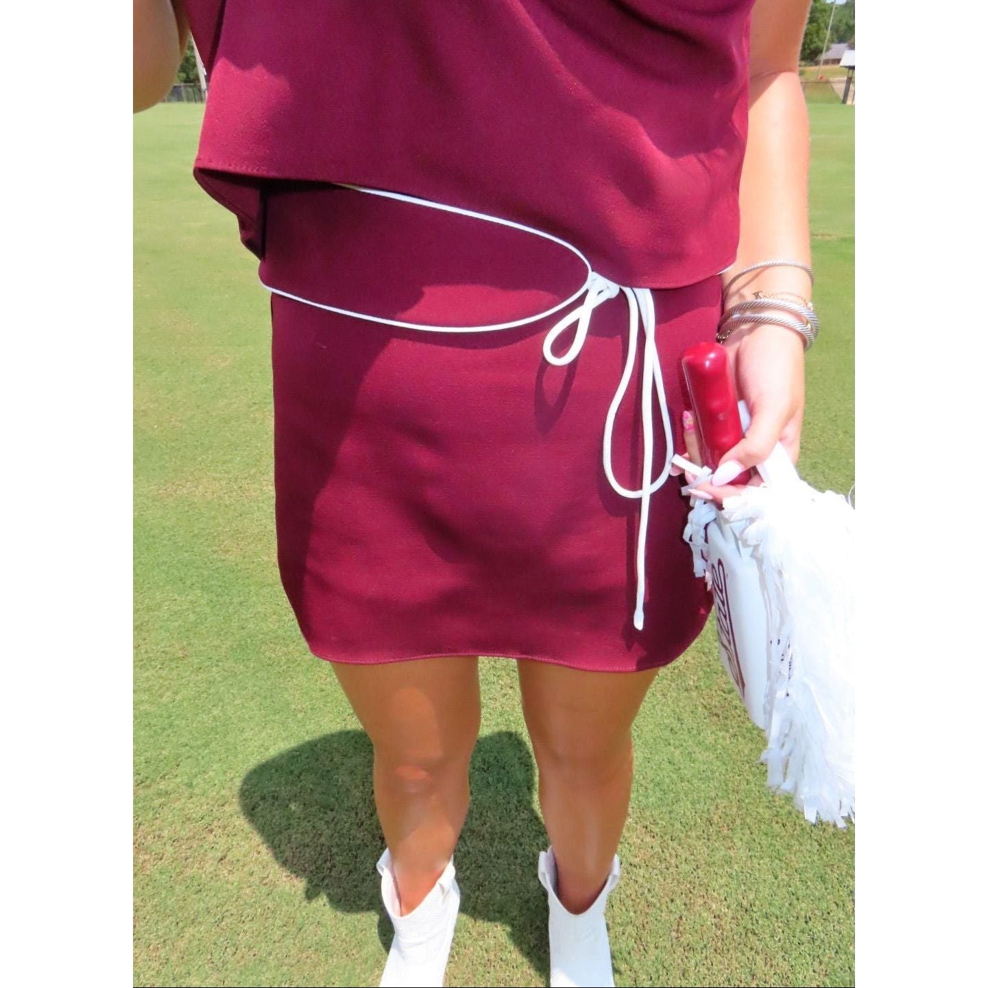 A person outdoors on a grassy lawn wearing a short burgundy dress with a white drawstring waist, holding a white paddle and a red phone.