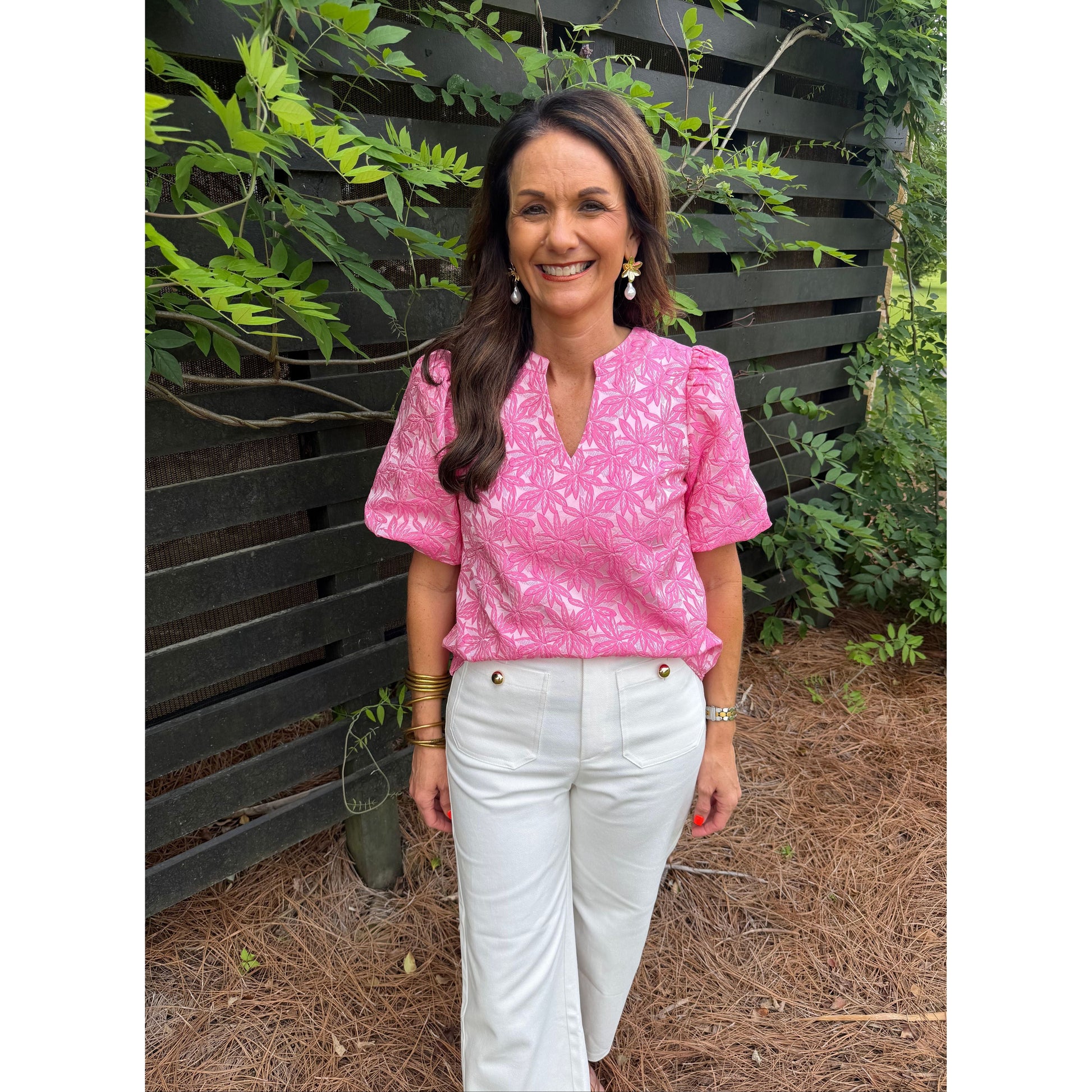 Woman in pink shirt and white pants standing outdoors with plants and a bench in the background