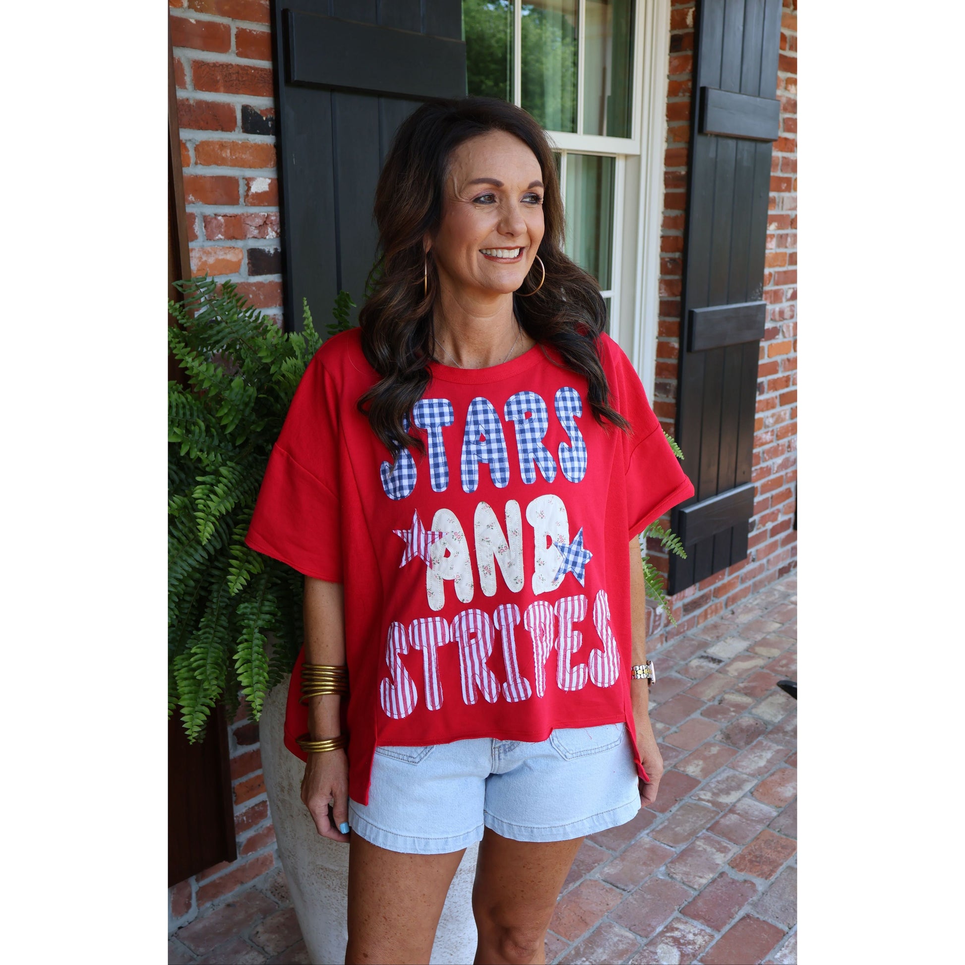 Woman wearing a red t-shirt with 'STARS AND STRIPES' text, standing outside a brick building.