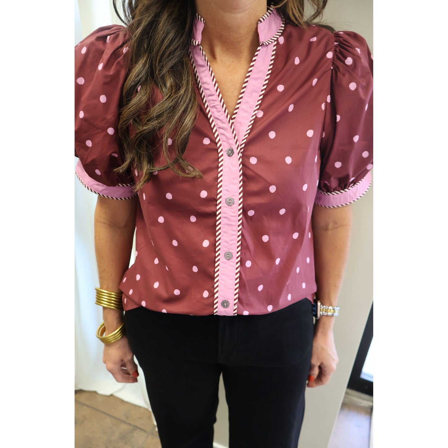 A woman wearing a dusty rose pink short-sleeve blouse with white polka dots, featuring a pink button-up front with striped trim and puff sleeves.