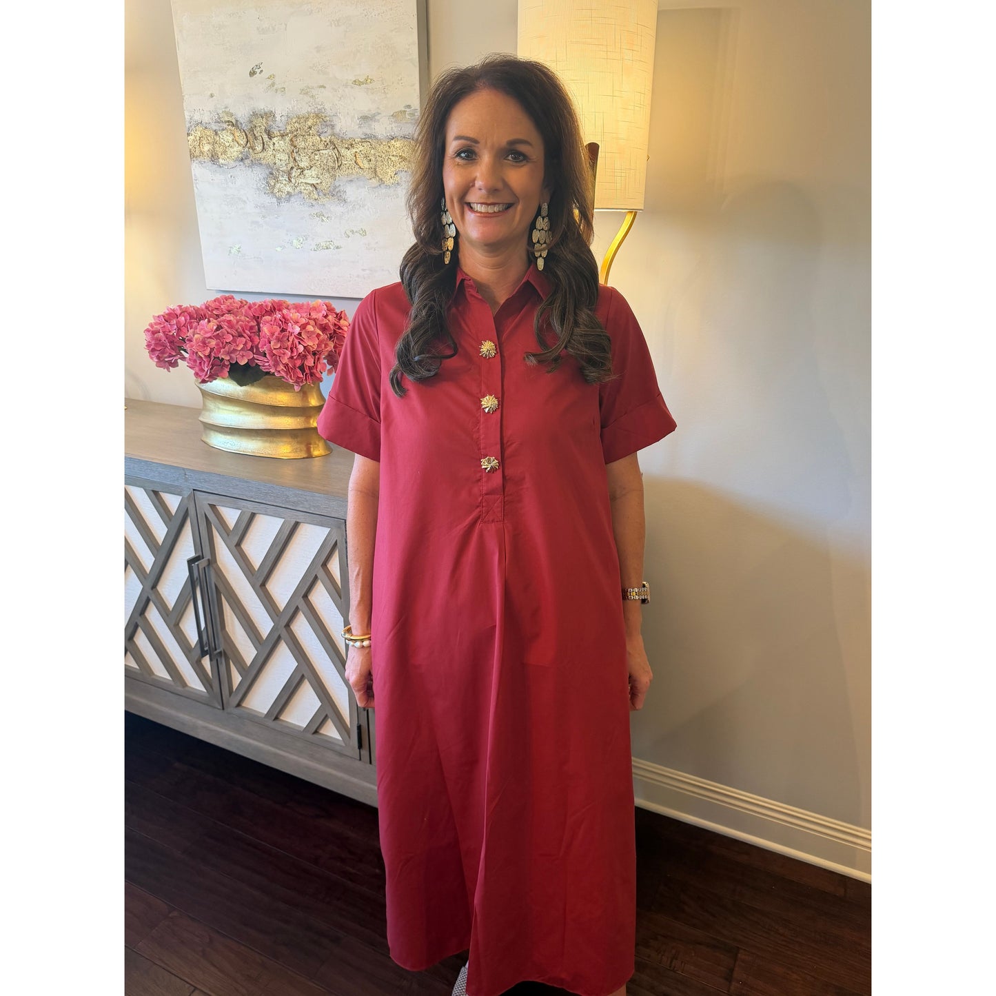 A woman wearing a short-sleeve red button-up dress with gold buttons, standing in a home interior setting.