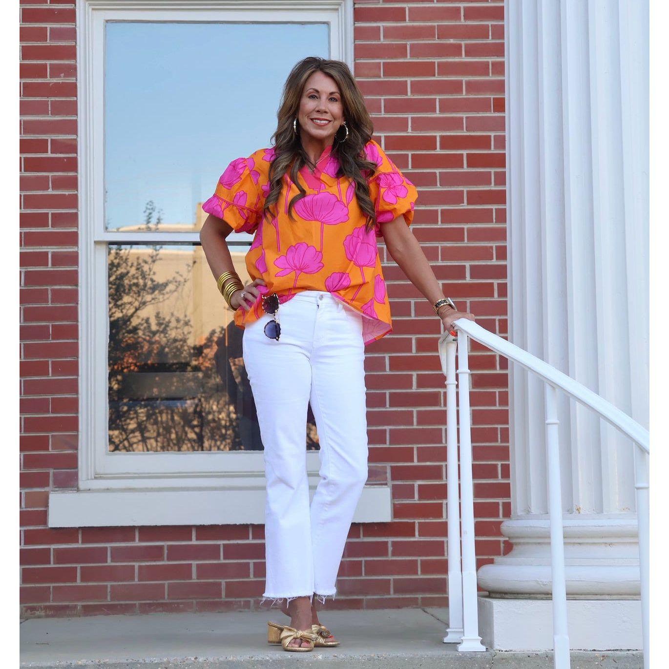 Woman in a colorful top and white pants standing in front of a brick building.