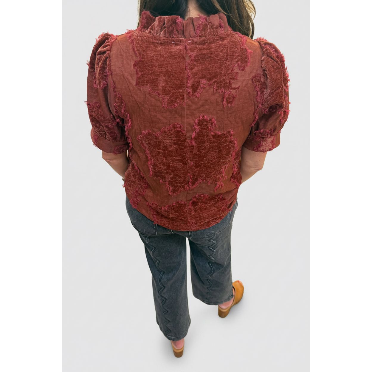 A person viewed from behind wearing a rust-red textured blouse with puff sleeves and a ruffled neckline, paired with dark gray jeans and tan shoes, standing in an indoor hallway.