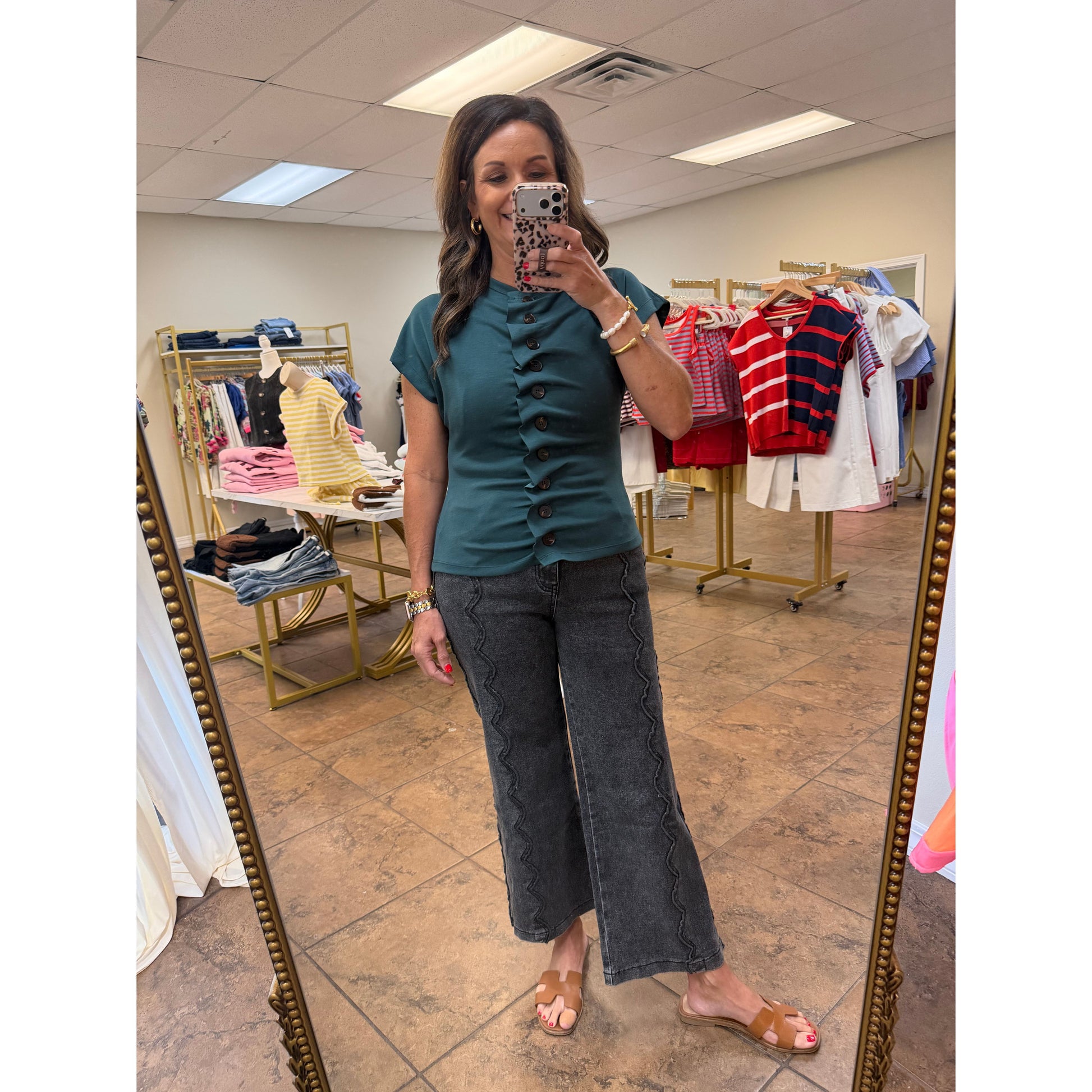A woman takes a mirror selfie in a boutique wearing a teal short-sleeve top with a button-down front and dark gray wide-leg pants. Clothing racks and store fixtures are visible in the background.