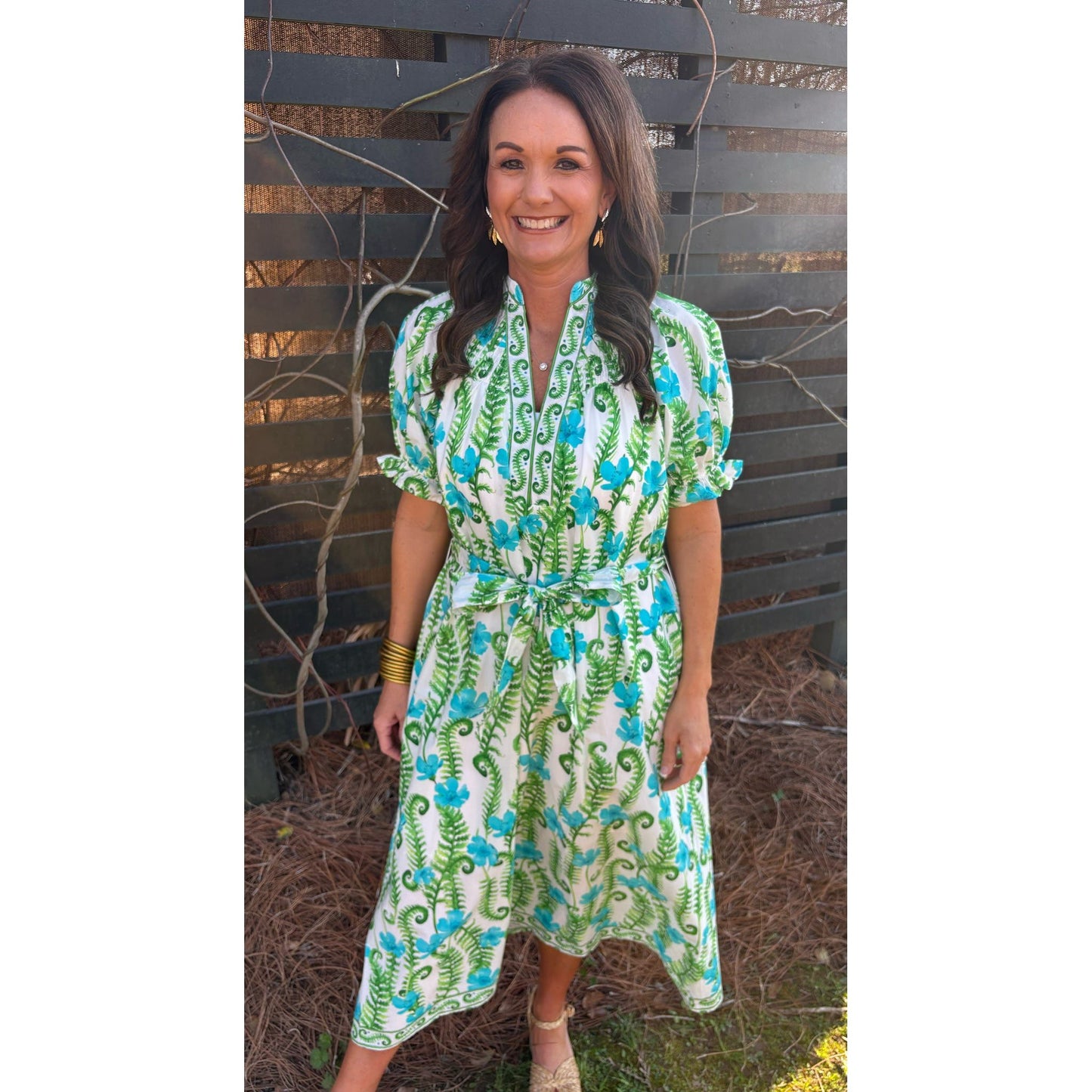 Woman wearing a green and blue patterned dress standing against a wooden fence.