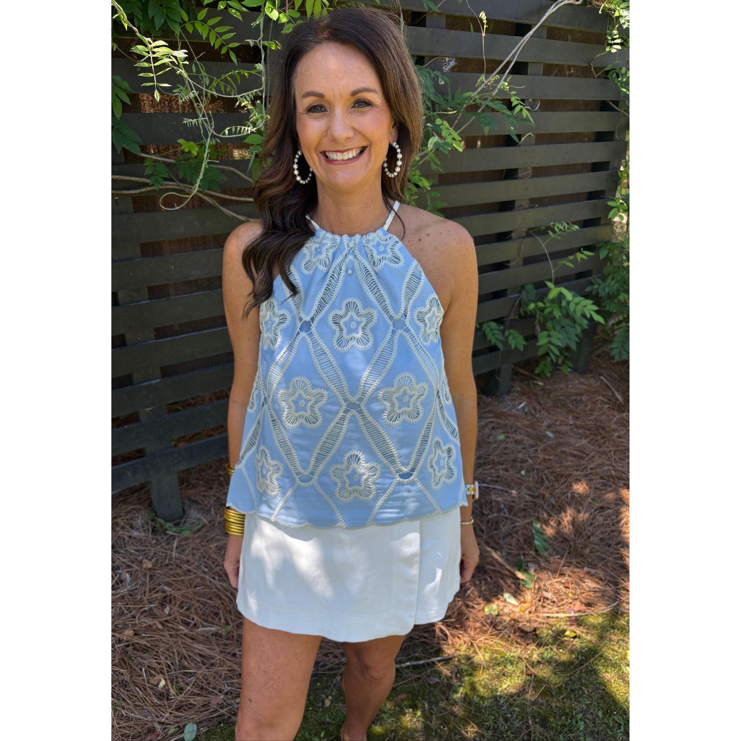 Woman wearing a blue patterned top and white shorts standing outdoors with greenery in the background