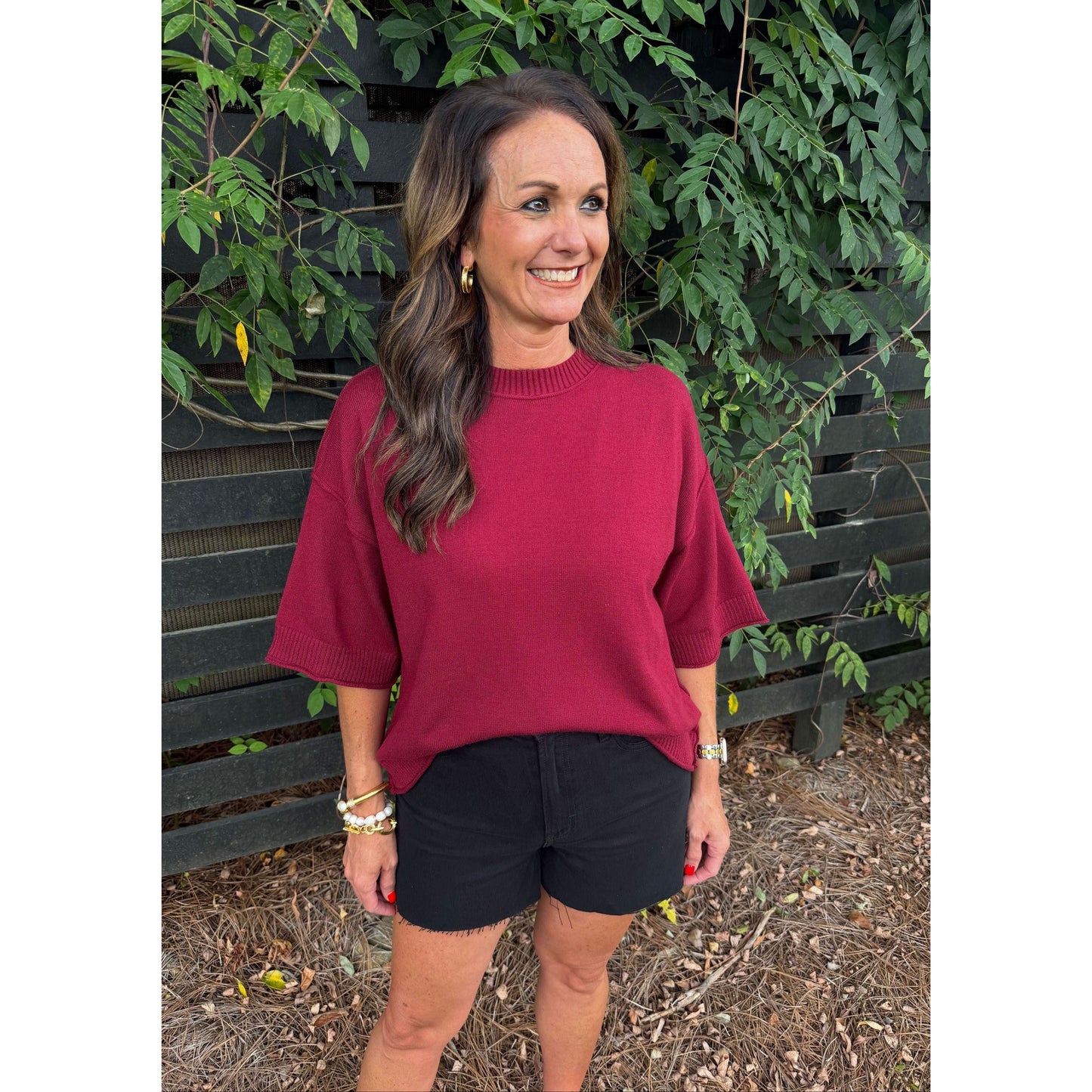 A woman wearing a burgundy short-sleeve knit top and black shorts, standing outdoors in front of a dark wooden fence and green foliage.
