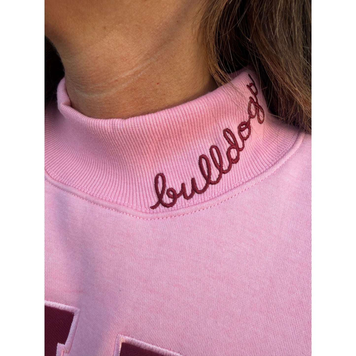 Close-up of a light pink sweatshirt with dark burgundy cursive embroidery on the ribbed collar, worn by a person.