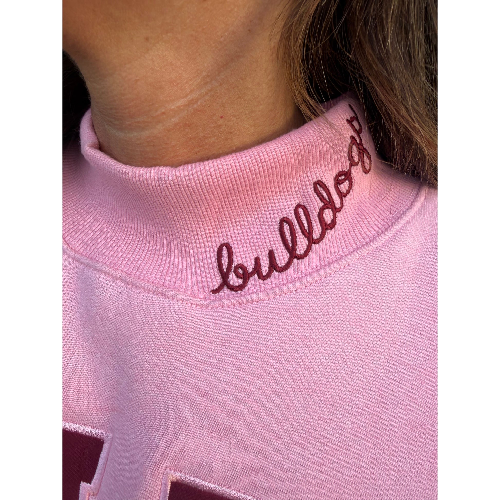 Close-up of a light pink sweatshirt with dark burgundy cursive embroidery on the ribbed collar, worn by a person.