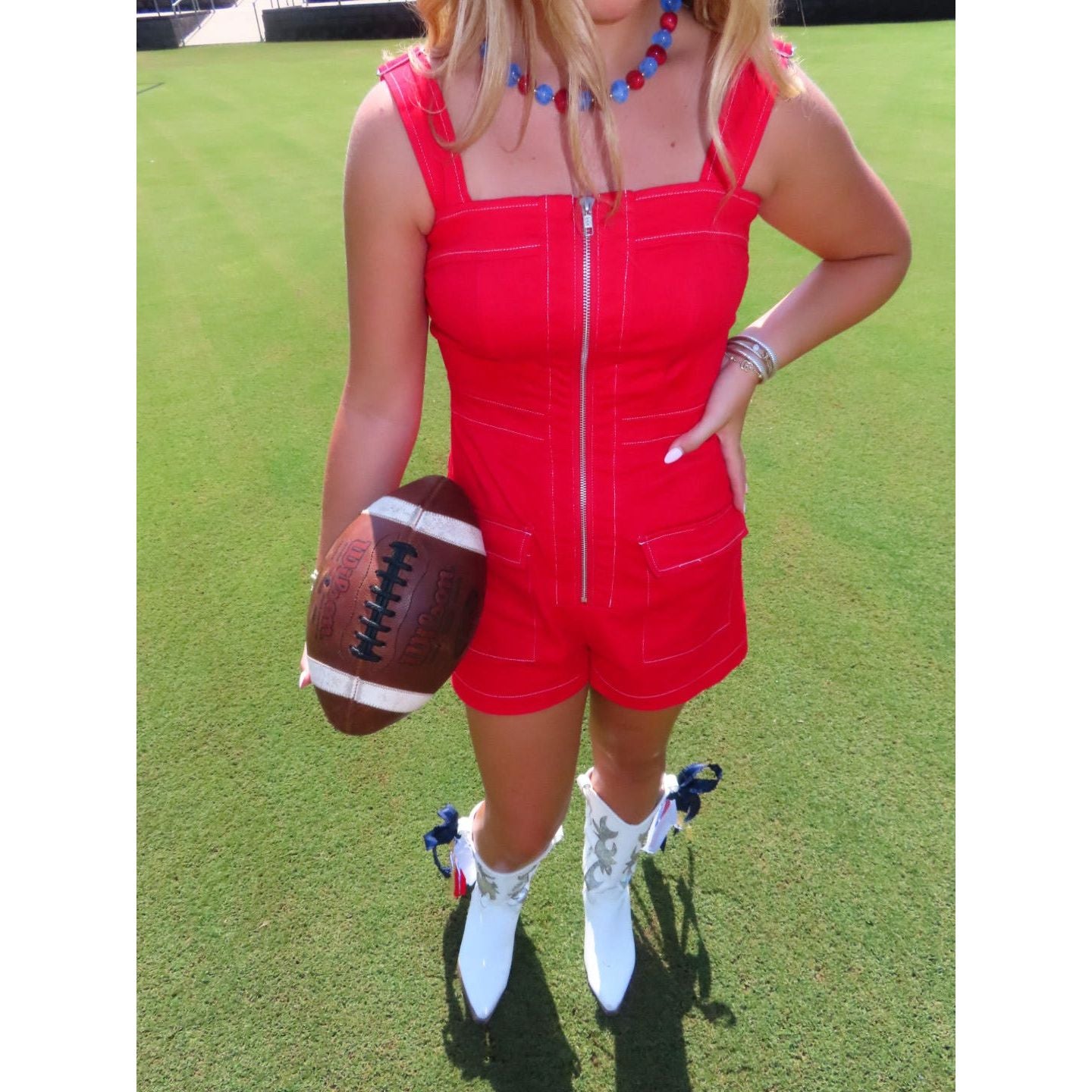 A woman standing on a grassy field wearing a bright red sleeveless romper with a front zipper and white western-style boots, while holding a brown football.