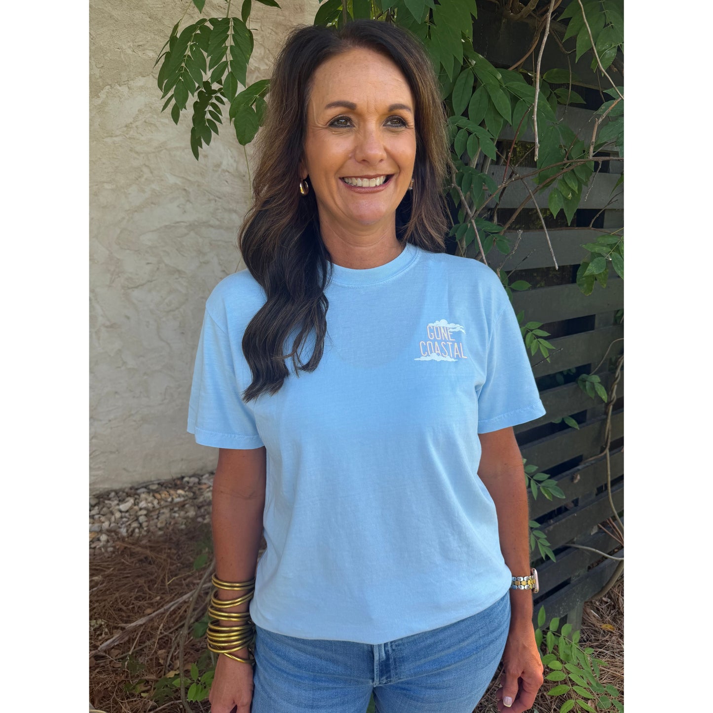 Woman wearing a light blue t-shirt with a logo, standing outdoors with greenery around.