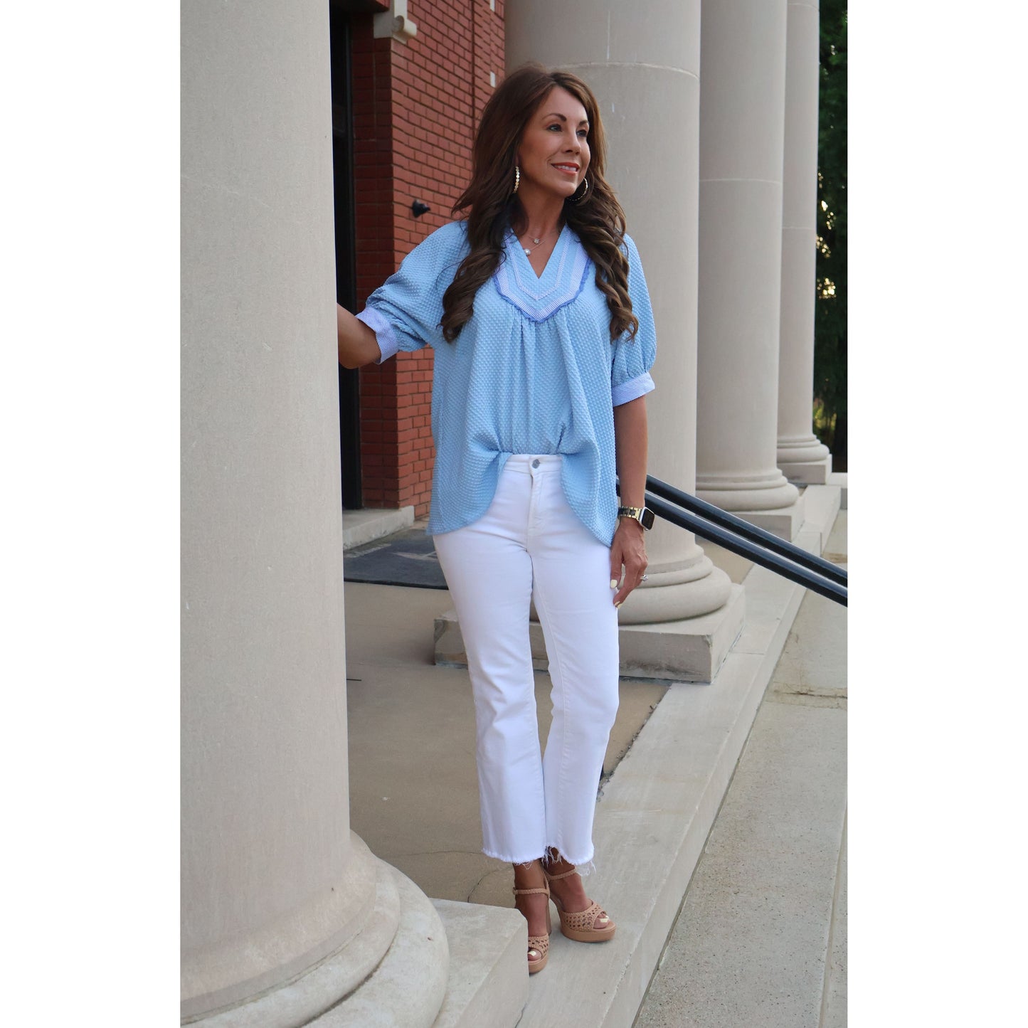 Woman in light blue top and white pants standing outdoors near a building entrance.