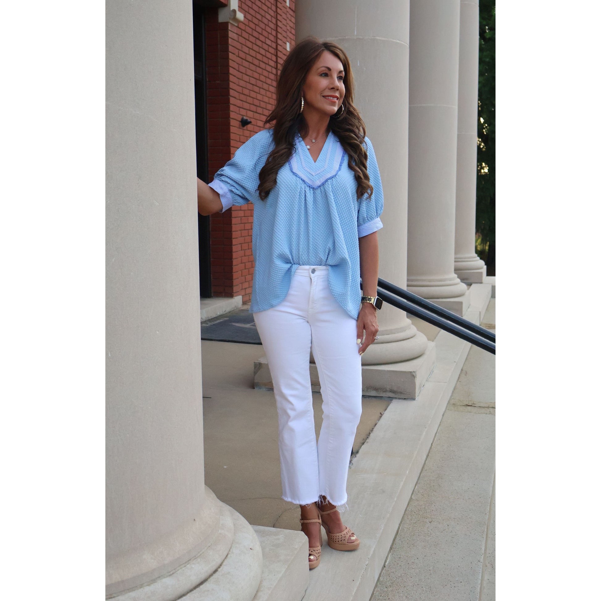Woman in light blue top and white pants standing outdoors near a building entrance.